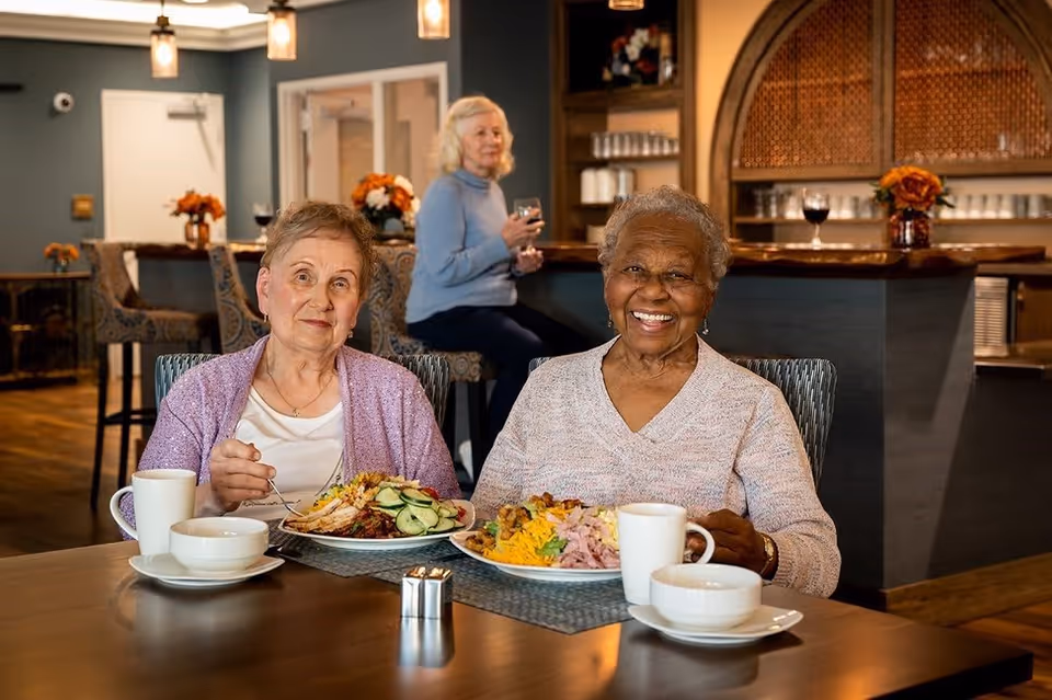 Two older women smiling at a dining table with plates of food in a cozy dining room while another woman stands at a bar in the background.