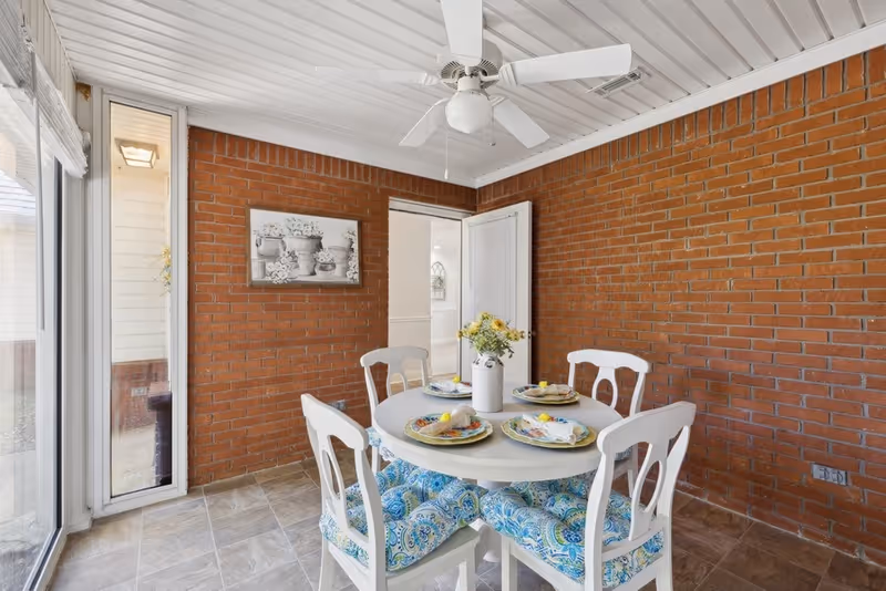 A bright dining area with a round white table set for four, featuring colorful plates and napkins. Four white chairs with blue patterned cushions surround the table. The room has a brick wall, a ceiling fan, and large windows letting in natural light.