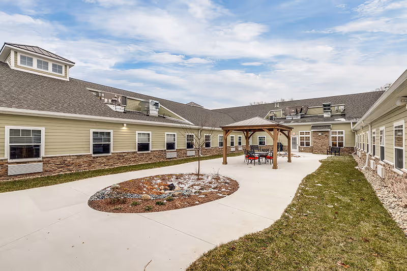 Outdoor courtyard area of a senior living facility with a paved walkway, a small garden bed with rocks and a young tree, and a wooden gazebo with a table and chairs underneath. The building surrounding the courtyard has light green siding with stone accents and multiple windows. The sky is partly cloudy.