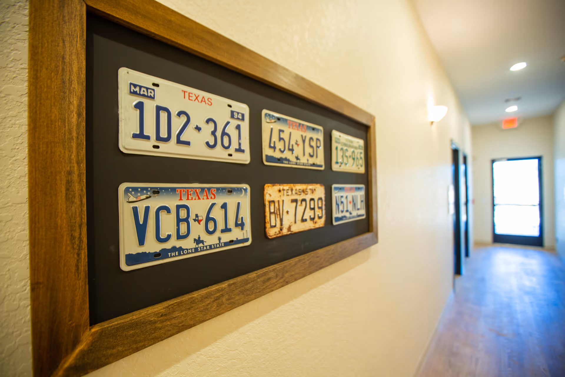 A framed display of six vintage Texas license plates mounted on a beige wall in a hallway with wooden flooring and a door with glass panels at the end.
