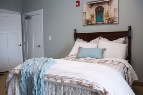 A neatly made bed with white and beige bedding, a light blue throw blanket, and a decorative pillow in a bedroom. The bed has a dark wooden headboard. On the wall above the bed is a framed picture of a green door with plants on either side. A white door and a closet door are visible in the background.