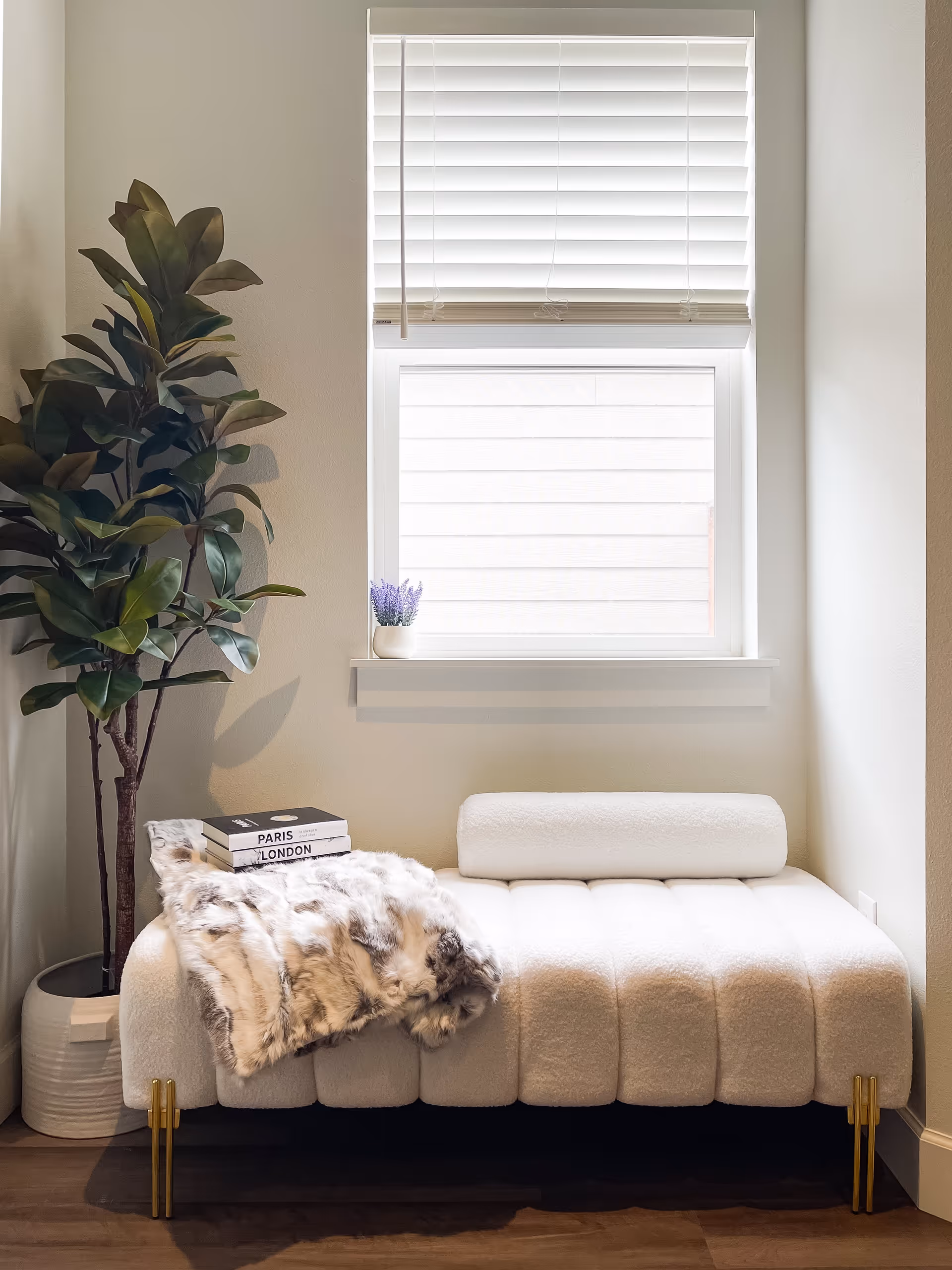 A cozy corner with a white cushioned bench featuring gold legs, a faux fur throw blanket, and two books titled 'PARIS' and 'LONDON' placed on the bench. Next to the bench is a tall potted plant with large green leaves. Above the bench is a window with white blinds partially closed, and a small white pot with purple flowers sits on the windowsill.