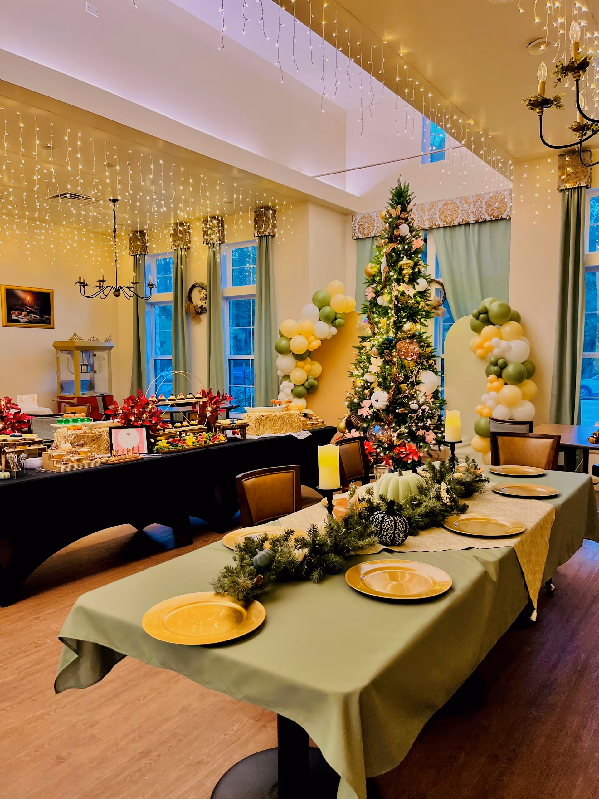 Festively decorated dining room with a Christmas tree, tables set with gold plates and garland centerpieces, a buffet table, string lights, and balloon arrangements.