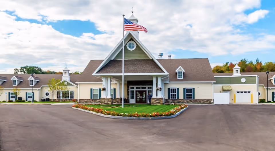 Front exterior view of Peregrine Senior Living at Onondaga Hill, a single-story building with a peaked roof, an American flag on a flagpole in front, and a landscaped flower bed surrounding the flagpole. The sky is partly cloudy.