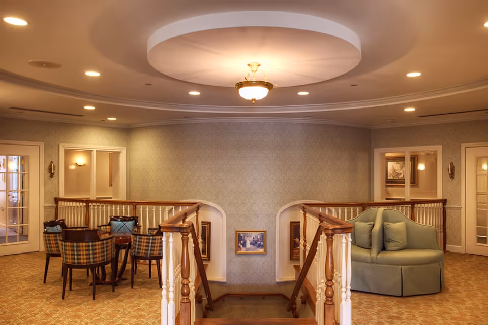 Upstairs common lounge with a staircase, round upholstered seat, table and chairs under recessed ceiling lighting.