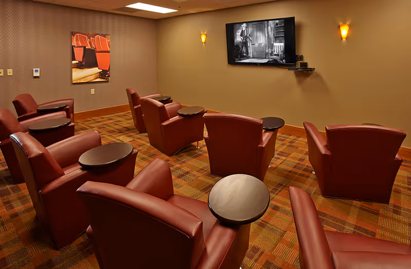 A cozy media room with several red leather recliner chairs arranged in rows facing a wall-mounted flat-screen TV playing a black and white movie. The room has warm lighting with two wall sconces and a patterned carpet in shades of brown and orange. A framed picture of theater seats hangs on one wall.