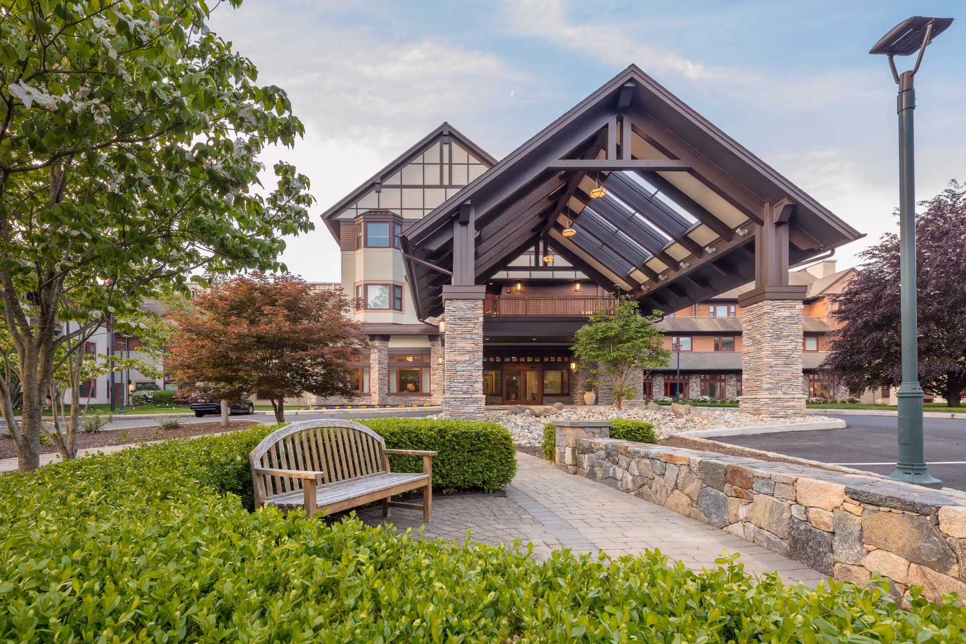 Entrance of Edgehill Senior Living Community Stamford featuring a large covered driveway with stone pillars, surrounded by well-maintained greenery including bushes, trees, and a wooden bench, with a stone wall and a streetlamp nearby under a partly cloudy sky.