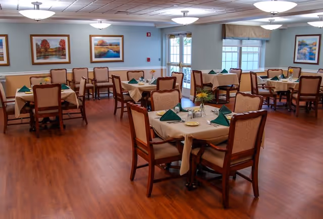 A dining room in a senior living facility with several tables covered in beige tablecloths, each set with green folded napkins, plates, and silverware. The room has wooden flooring, cushioned chairs, framed landscape paintings on the walls, and large windows letting in natural light.