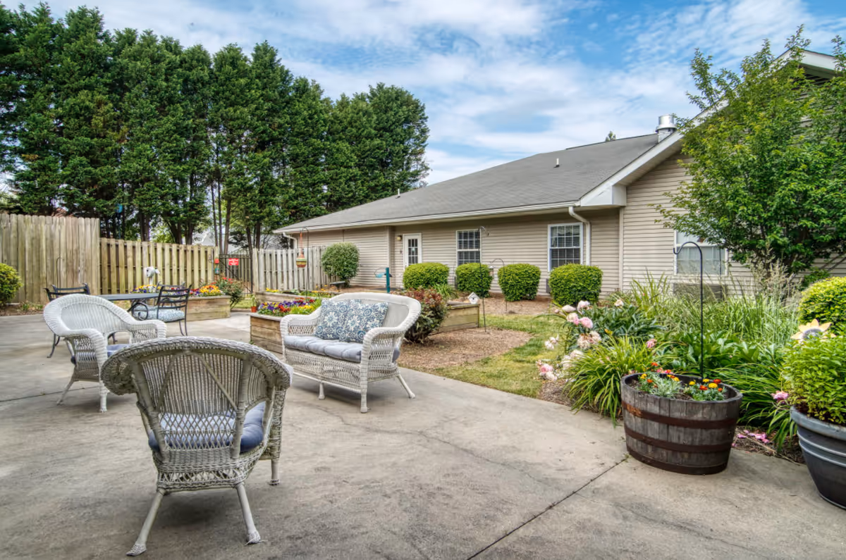 Outdoor patio courtyard with white wicker chairs and a loveseat, potted flowers and garden beds in front of a single-story building.