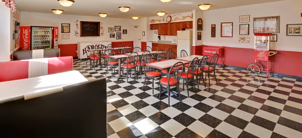 A retro-style dining room with black and white checkered flooring, red and black chairs, and red and white booths. The walls are painted white and red, decorated with framed pictures and a chalkboard sign that reads 'Harold's Deli'. There is a Coca-Cola vending machine and a popcorn machine in the room, along with a kitchenette area with wooden cabinets, a refrigerator, and a coffee maker.