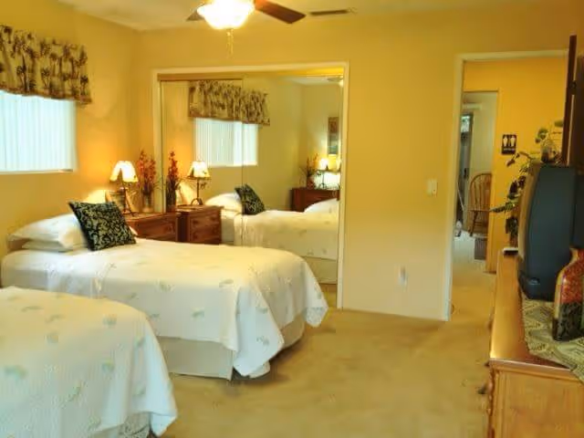 A bedroom with two single beds covered in white bedspreads with subtle patterns. Each bed has a decorative pillow. There is a wooden nightstand with two lamps and floral decorations between the beds. A large mirrored closet door reflects the room. The walls are painted light yellow, and there is a window with a patterned valance. A wooden dresser with a TV and decorative items is visible on the right side.