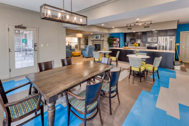 A spacious and modern common area in a senior living facility featuring a large wooden dining table with six chairs, smaller round tables with chairs, a kitchen area with dark cabinetry and stainless steel appliances, and a seating area with blue armchairs and a yellow ottoman. The floor has a blue and beige geometric pattern, and there is a glass door leading to an outdoor patio with metal chairs and tables.