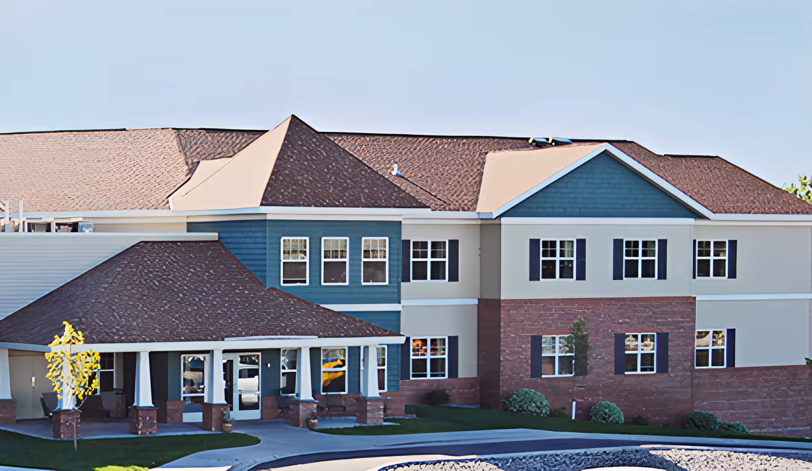 Exterior view of a two-story senior living facility building with a combination of blue siding, beige walls, and red brick accents. The building has multiple windows with black shutters and a covered entrance supported by white columns. The sky is clear and the surrounding area includes a driveway and some landscaping.