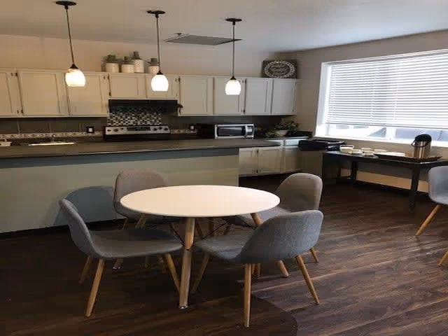 A modern kitchen and dining area with a round white table surrounded by four gray upholstered chairs with wooden legs. The kitchen features white cabinets, a stove, a microwave, and three pendant lights hanging from the ceiling. There is a large window with blinds letting in natural light, and a side table with coffee cups and a coffee maker.