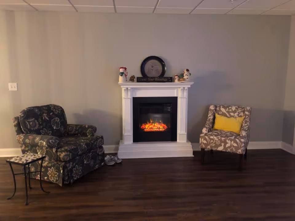A cozy living room area with a white electric fireplace in the center, decorated with small holiday figurines and a round clock on the mantel. To the left of the fireplace is a floral patterned armchair with a small side table, and to the right is a beige patterned chair with a yellow cushion. The room has wooden flooring and light gray walls.