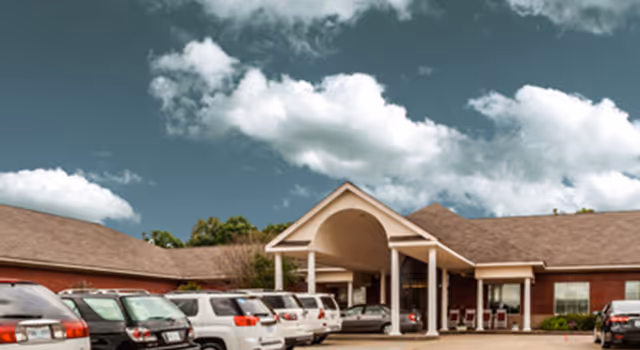 Front entrance of a brick senior living building with a covered porte-cochere and several parked cars under a cloudy sky.