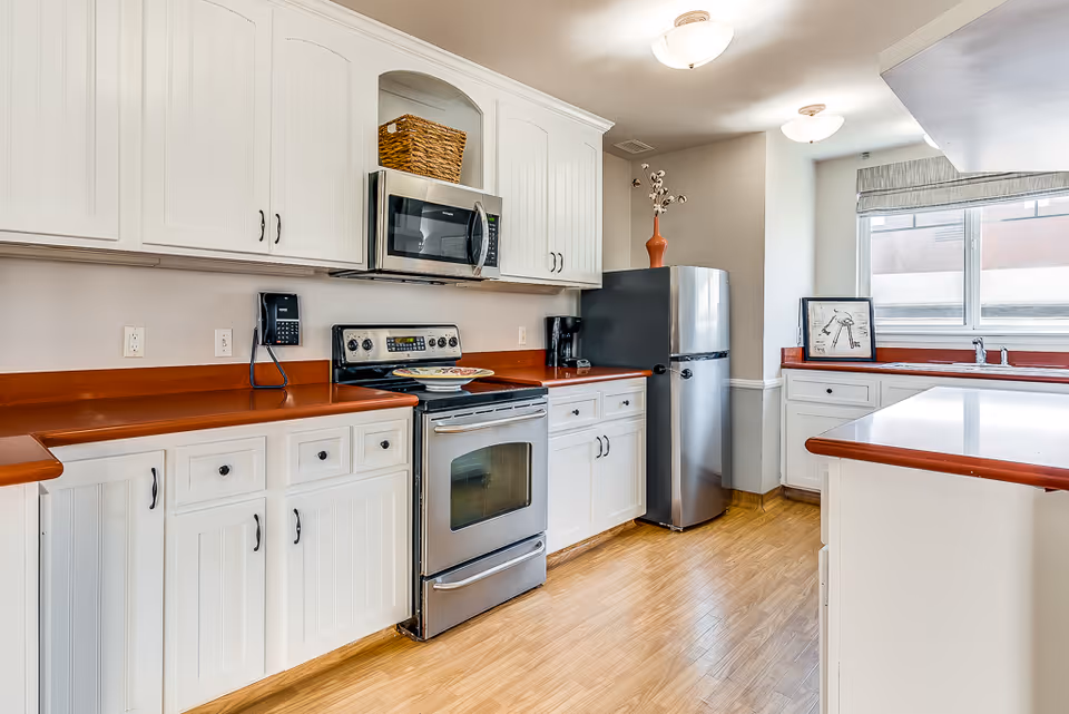 A bright kitchen with white cabinets and red countertops. The kitchen features a stainless steel oven with a microwave above it, a stainless steel refrigerator, a coffee maker, and a window letting in natural light. The floor is wooden, and there is a decorative basket on top of the cabinets and a vase with flowers on the refrigerator.