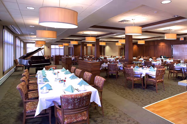 Large dining room with multiple set tables and chairs, pendant lights, folded napkins, and a grand piano by the windows.