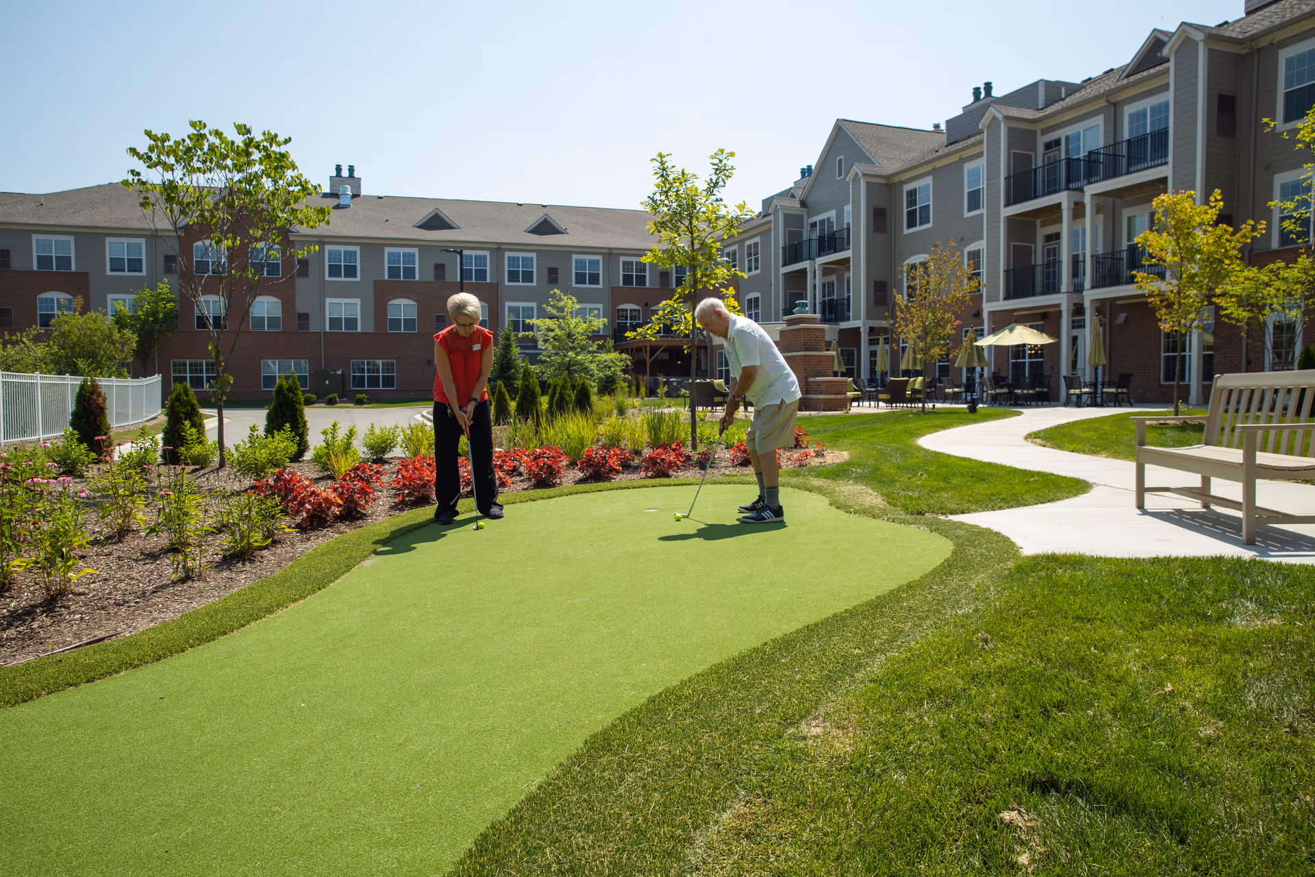 Two elderly people playing mini golf on a putting green in the garden area of a senior living facility. The background shows a multi-story residential building with balconies, trees, shrubs, and outdoor seating areas under umbrellas.