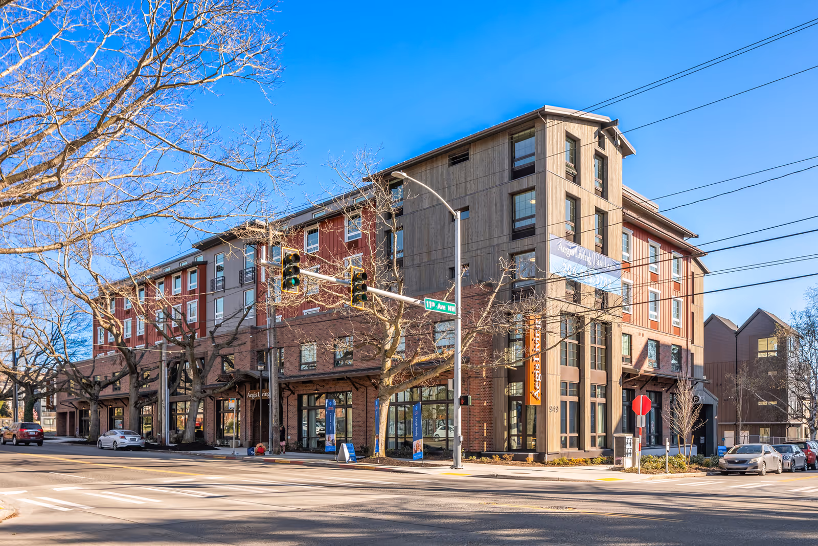 Corner view of a multi-story Aegis Living Ballard senior living building with storefronts, trees, and parked cars under a clear blue sky.