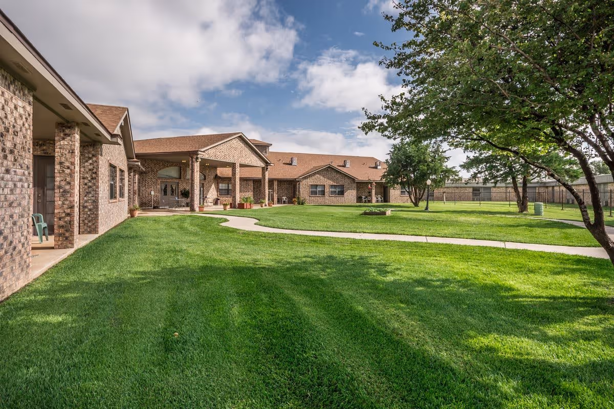 A well-maintained outdoor courtyard area of a senior living facility with green grass, a paved walkway, several trees, and a brick building with covered porch areas under a partly cloudy sky.
