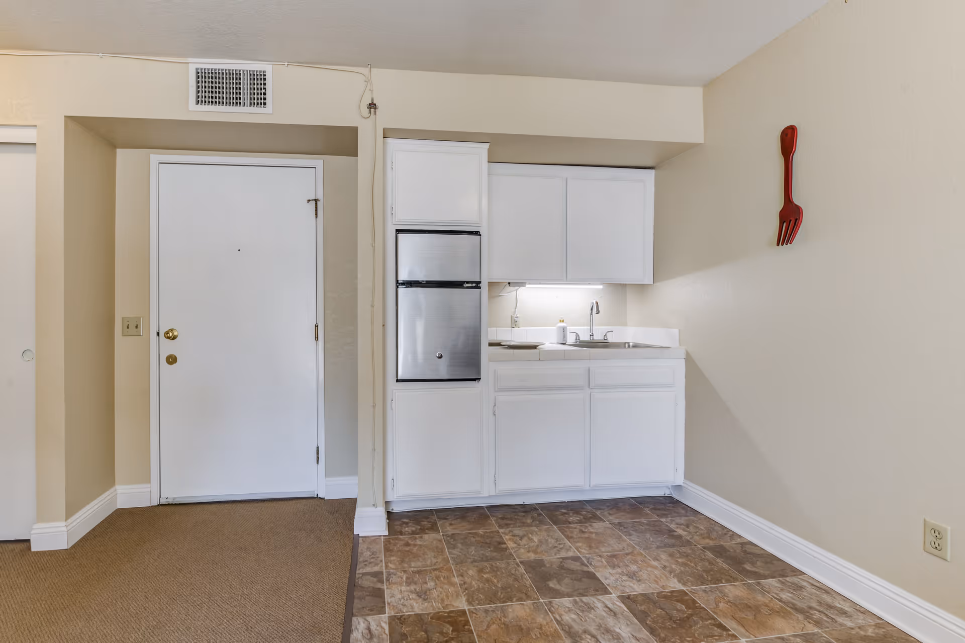 Small kitchenette area with white cabinets, a stainless steel mini refrigerator, a sink, and a countertop. To the left is a white door and beige walls. The floor is a combination of brown carpet and brown tile. A large red decorative fork hangs on the wall to the right.