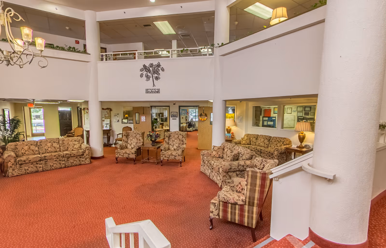 Spacious two-story senior living lobby with patterned sofas and armchairs arranged on a red carpet under an open balcony.