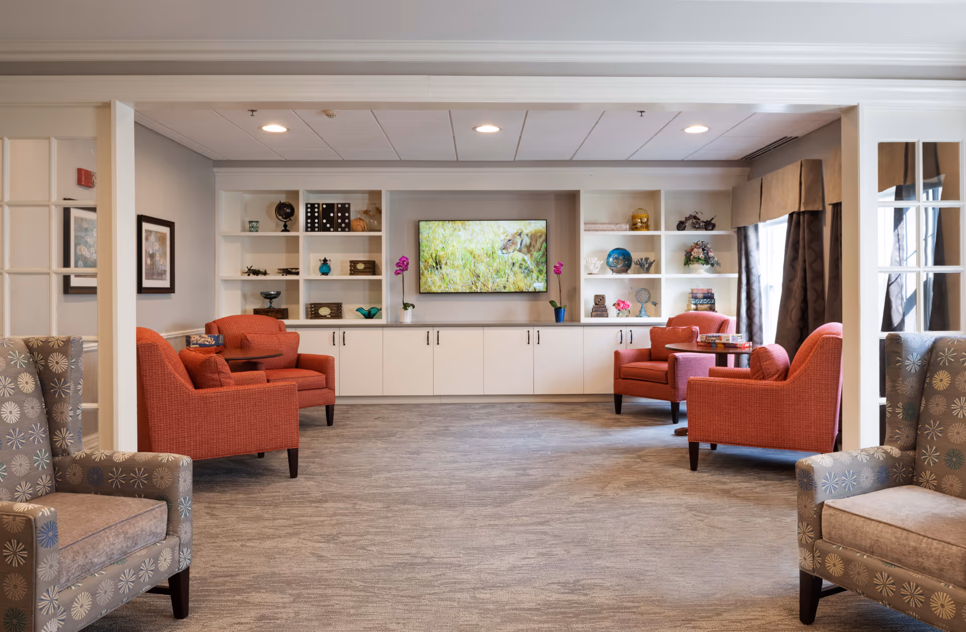 A cozy senior living common area with four red armchairs and two patterned gray armchairs arranged around small tables. Built-in white shelves and cabinets line the back wall, decorated with various ornaments and a flat-screen TV displaying a nature scene. Large windows with brown curtains allow natural light into the room.