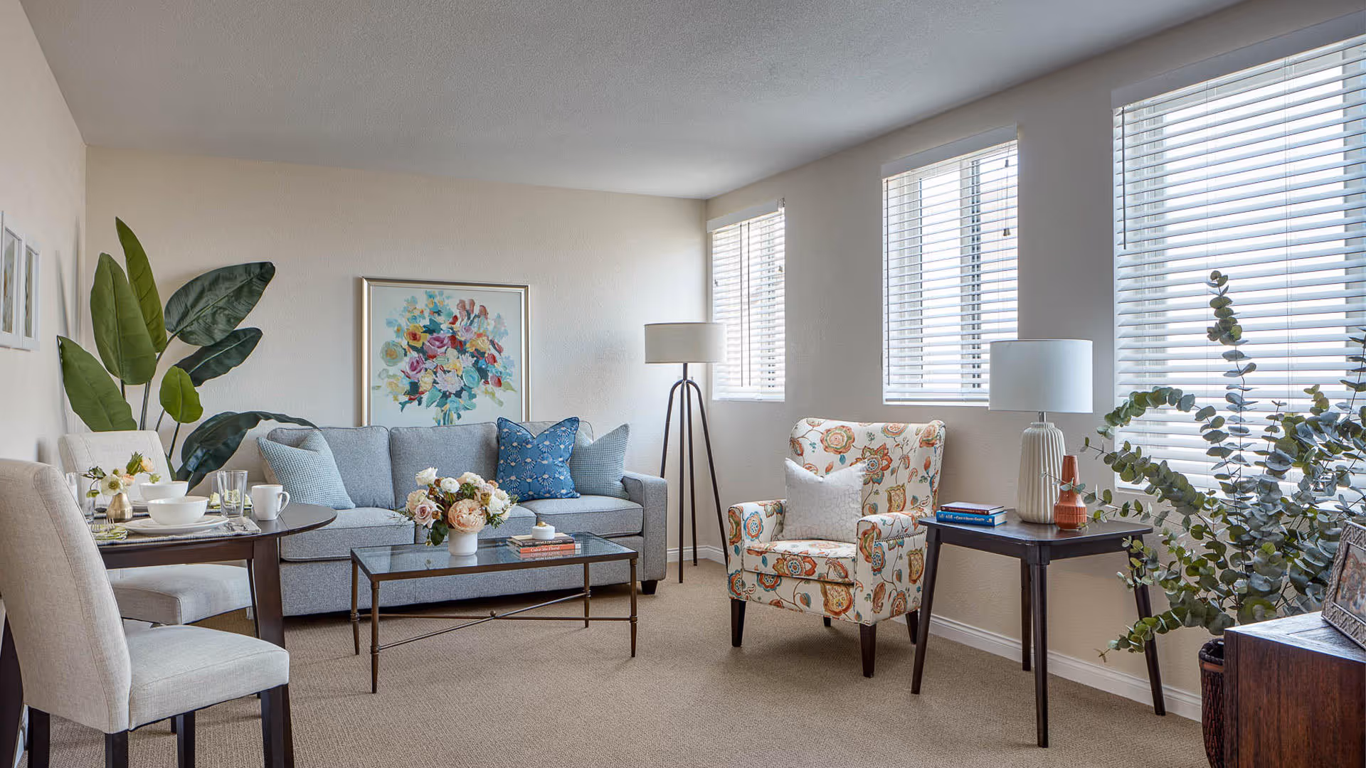 A bright and cozy living room with a light gray sofa adorned with blue and patterned pillows, a floral armchair, a glass coffee table with a flower arrangement, a round dining table set with dishes and glasses, two side tables with lamps, large green plants, and three windows with white blinds letting in natural light.