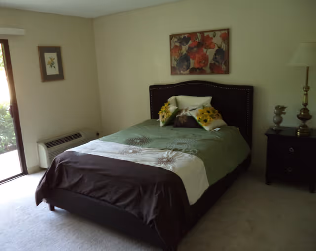 A bedroom with a neatly made bed featuring a green and brown bedspread and decorative pillows with sunflower designs. There is a nightstand with a lamp and decorative items on the right side of the bed. A framed floral painting hangs above the headboard. A sliding glass door is visible on the left side, allowing natural light into the room.
