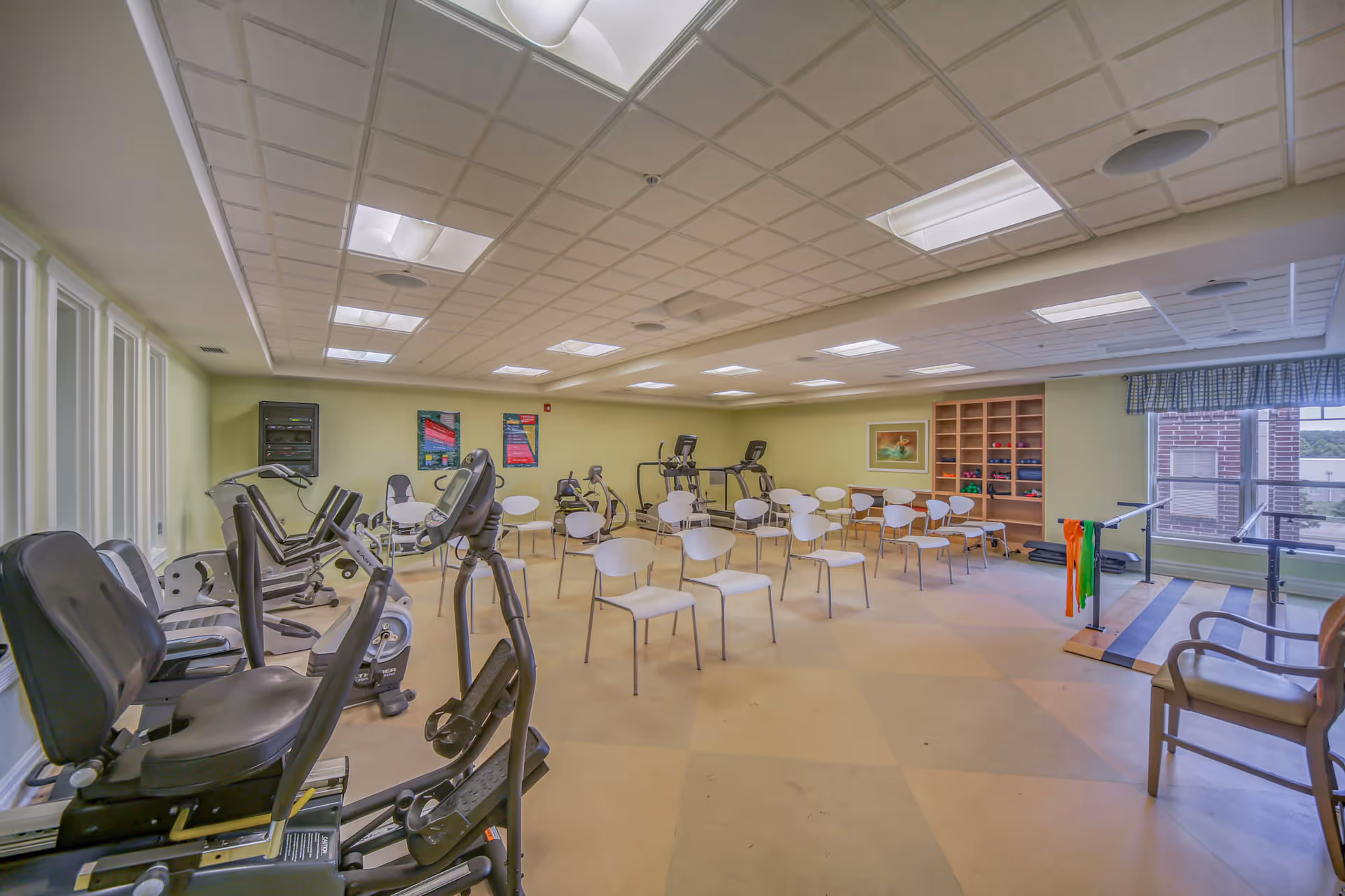 Bright fitness/activity room with multiple exercise machines, rows of white chairs, shelving, and large windows.
