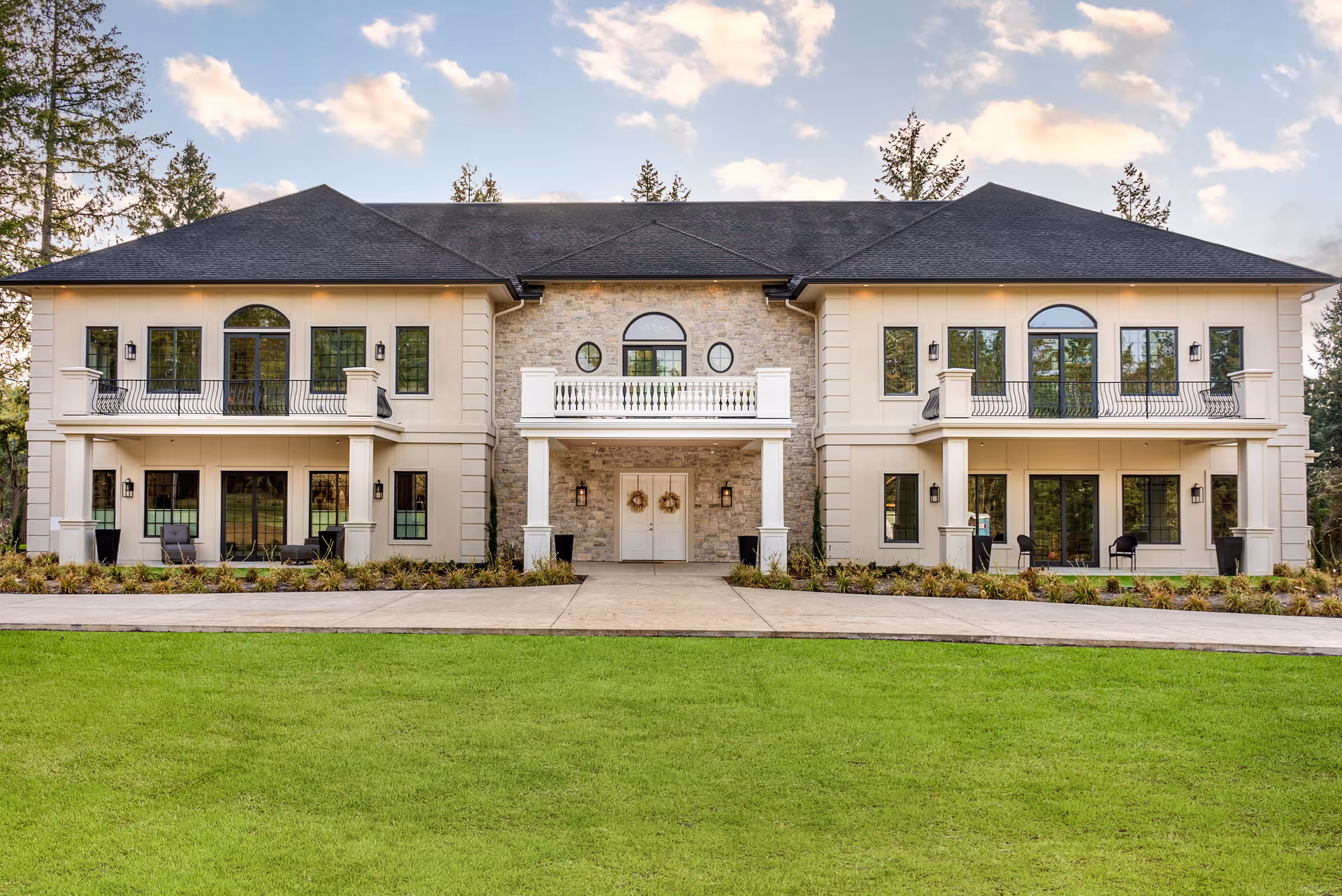 A two-story beige residential building with a central stone entry and balconies set behind a manicured green lawn.