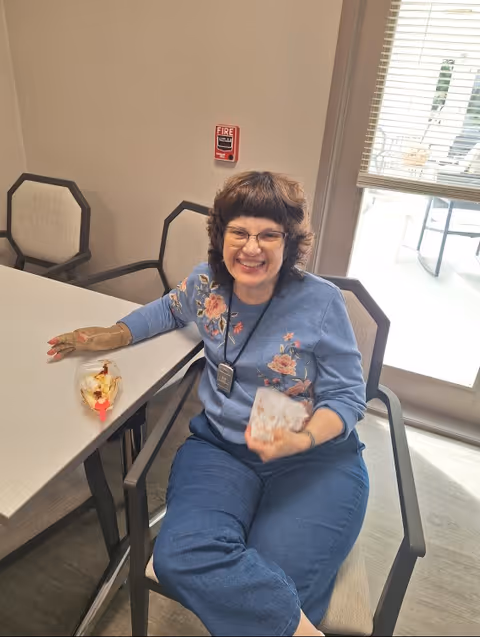 A smiling woman with glasses and curly hair sits at a table in a room with beige walls and chairs. She is wearing a blue floral sweater and blue pants, holding a card in one hand and resting the other on the table near a partially eaten dessert. Behind her is a window with blinds and a door leading outside.
