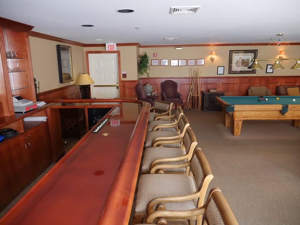 Interior view of a recreational room featuring a long wooden bar with several cushioned chairs lined up in front of it. In the background, there is a pool table with balls set up, a few armchairs, framed pictures on the wall, and a lamp on a side table. The room has wood paneling and a beige carpeted floor.