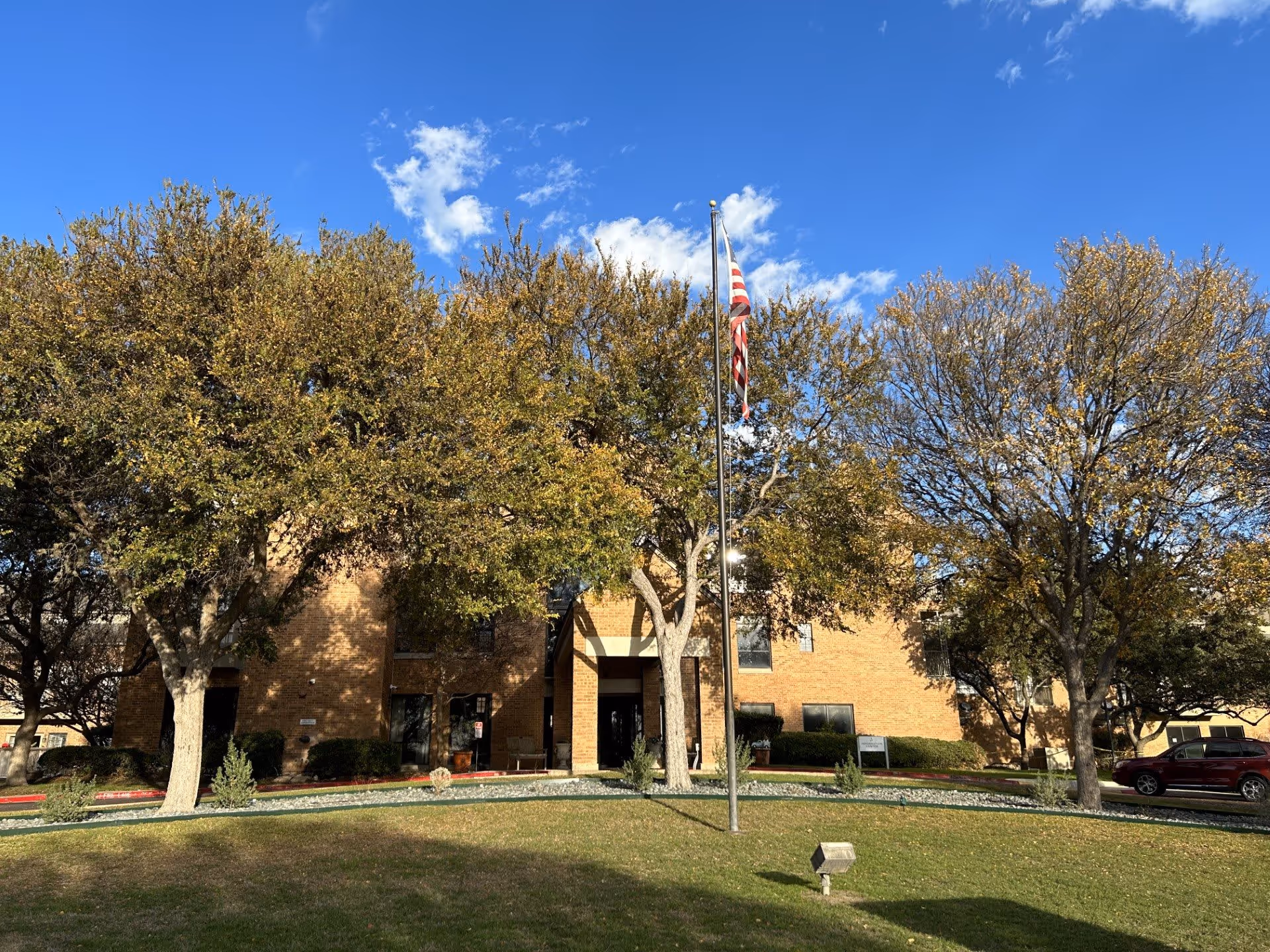 Exterior view of a brick building partially obscured by several trees with autumn foliage. In front of the building is a flagpole with an American flag. The sky is clear and blue with a few scattered clouds. There is a grassy lawn and a small spotlight in the foreground, and a red vehicle is parked to the right side near the building.