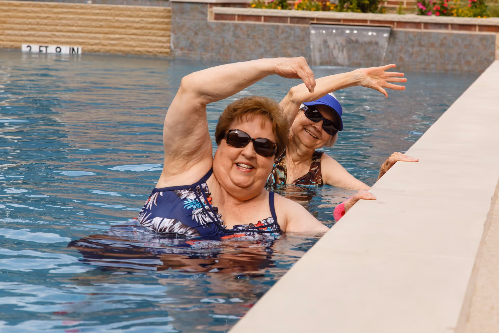 Two elderly women wearing sunglasses and swimsuits enjoying themselves in a swimming pool, smiling and posing with one arm raised while holding onto the pool edge.
