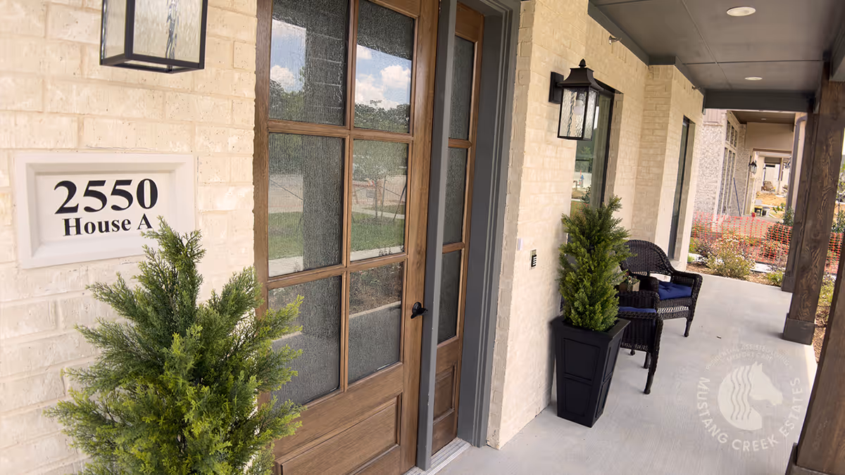 Front porch of a senior living facility unit with a wooden door featuring glass panels, two potted green plants, two black wicker chairs, and a sign on the wall that reads '2550 House A'. The porch has a covered ceiling with recessed lighting and wooden support beams.