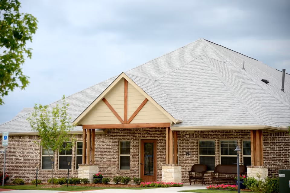 Exterior view of a single-story brick building with a light gray shingled roof, a wooden front porch with two chairs, small trees, and landscaped flower beds in front.