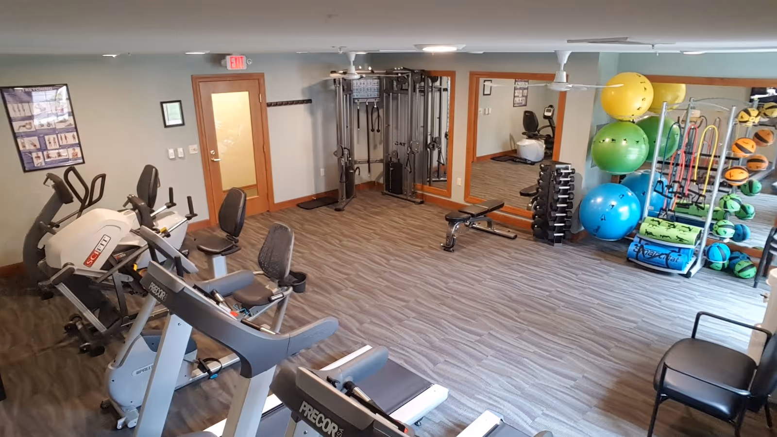 A fitness room with various exercise equipment including stationary bikes, a treadmill, weight bench, dumbbells, resistance bands, exercise balls, and medicine balls. The room has wood-patterned flooring, a large mirror on one wall, and a door with a glass panel. There are posters on the walls and a chair in the corner.