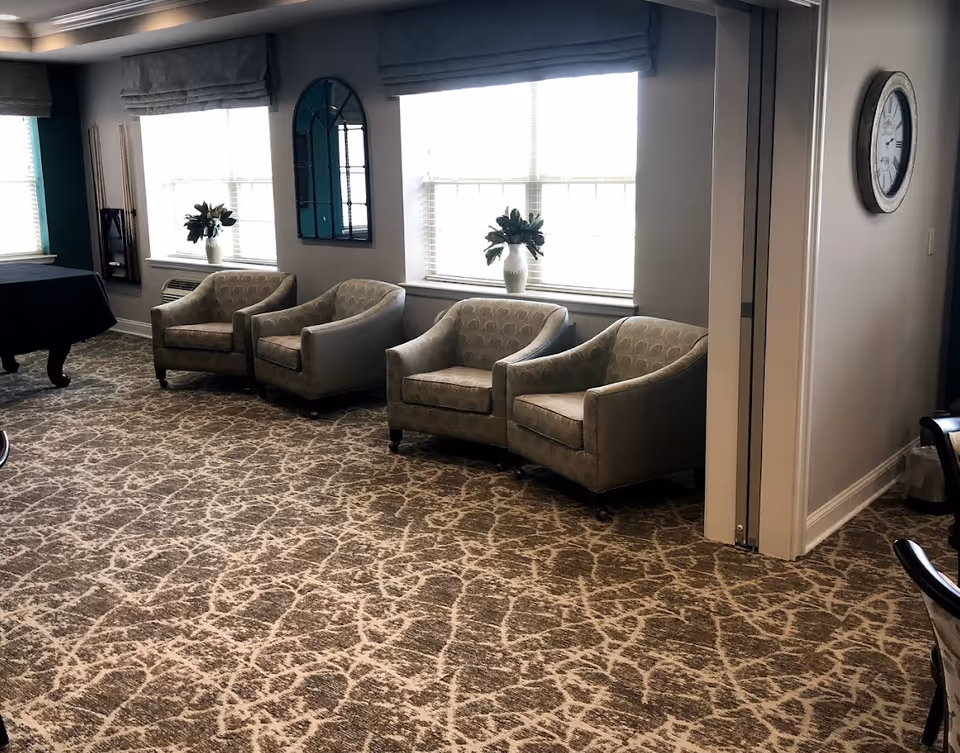 A sitting area in Avalon Assisted Living at Hillsborough featuring four beige armchairs arranged in a row along a wall with two large windows. Each window has a vase with flowers on the windowsill. The room has patterned carpet flooring, a wall clock, and a pool table partially visible on the left side.
