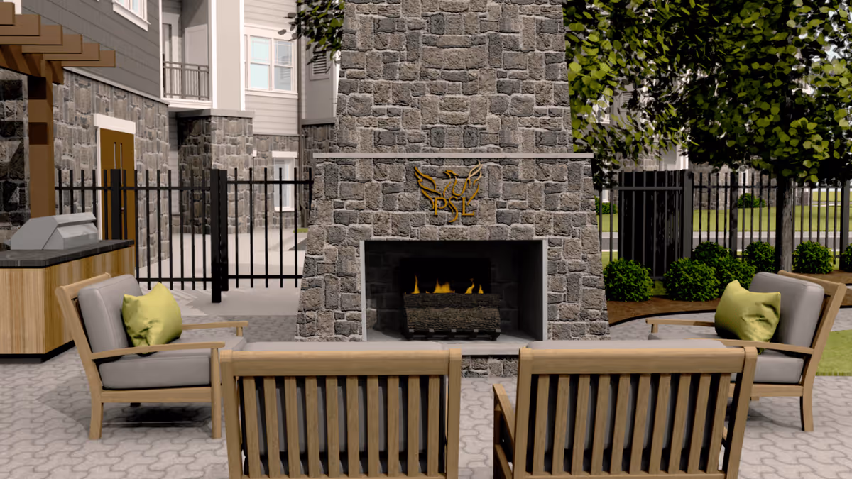 Outdoor patio area with a stone fireplace featuring a decorative emblem. The fireplace is surrounded by four cushioned wooden chairs with green pillows. There is a built-in grill station to the left and a black metal fence enclosing the area. Trees and shrubs are visible in the background.
