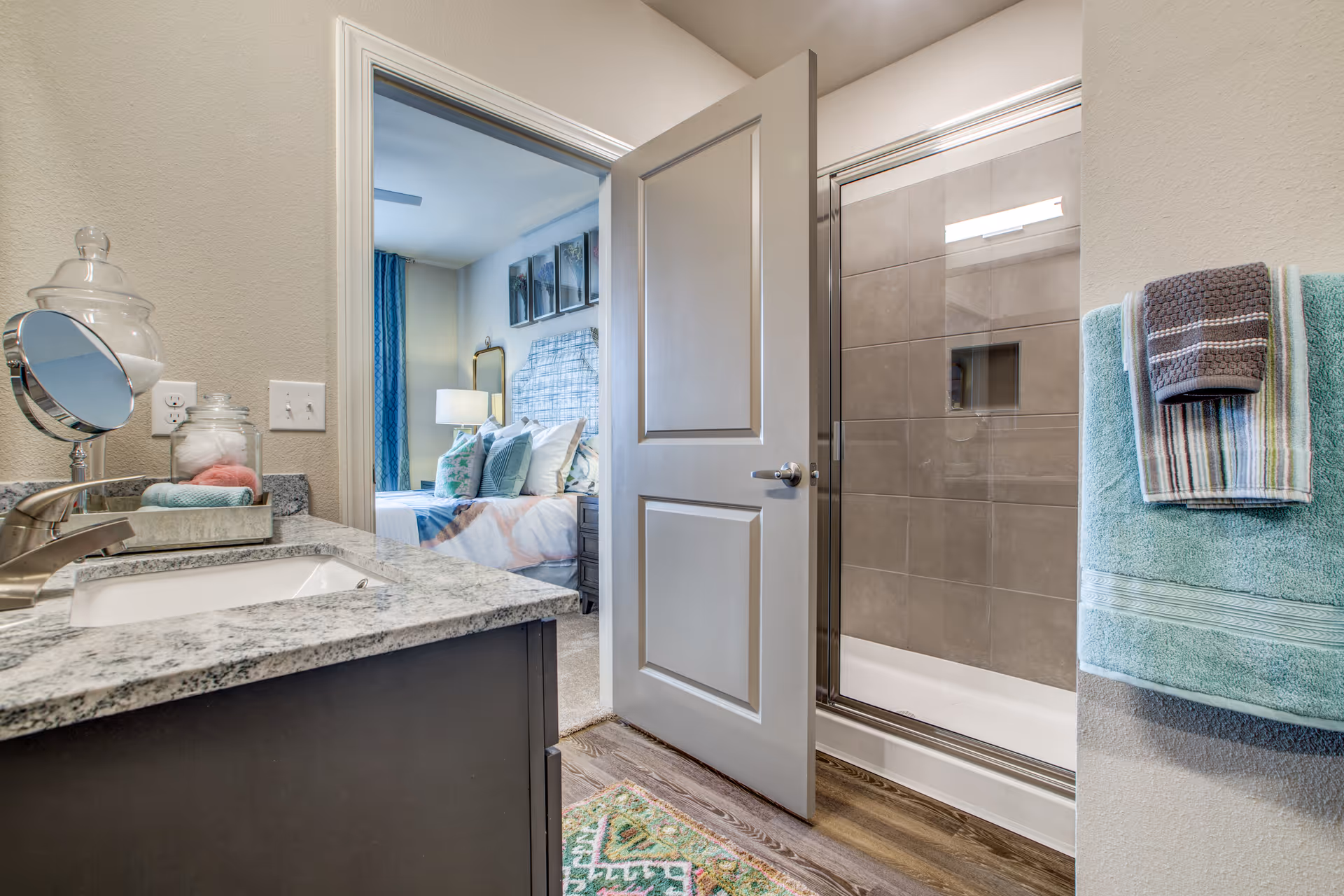 View of a bathroom with a granite countertop sink, a glass jar with cotton balls, and a small mirror on the left. The bathroom has a glass-enclosed shower with gray tiles and a towel rack holding folded towels on the right. The bathroom door is open, revealing a bedroom with a bed, pillows, a lamp, and wall art in the background.
