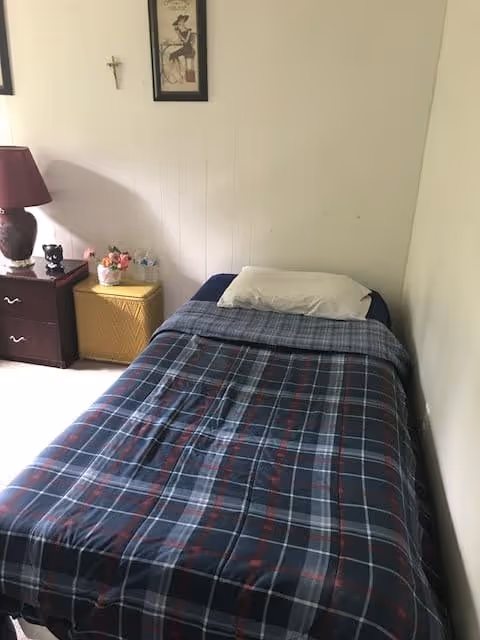 A small bedroom with a single bed covered in a blue and red plaid blanket and a white pillow. Next to the bed is a dark wooden nightstand with a red lamp, a small black figurine, and a basket with flowers and water bottles. A framed picture and a small cross hang on the white wall above the nightstand.