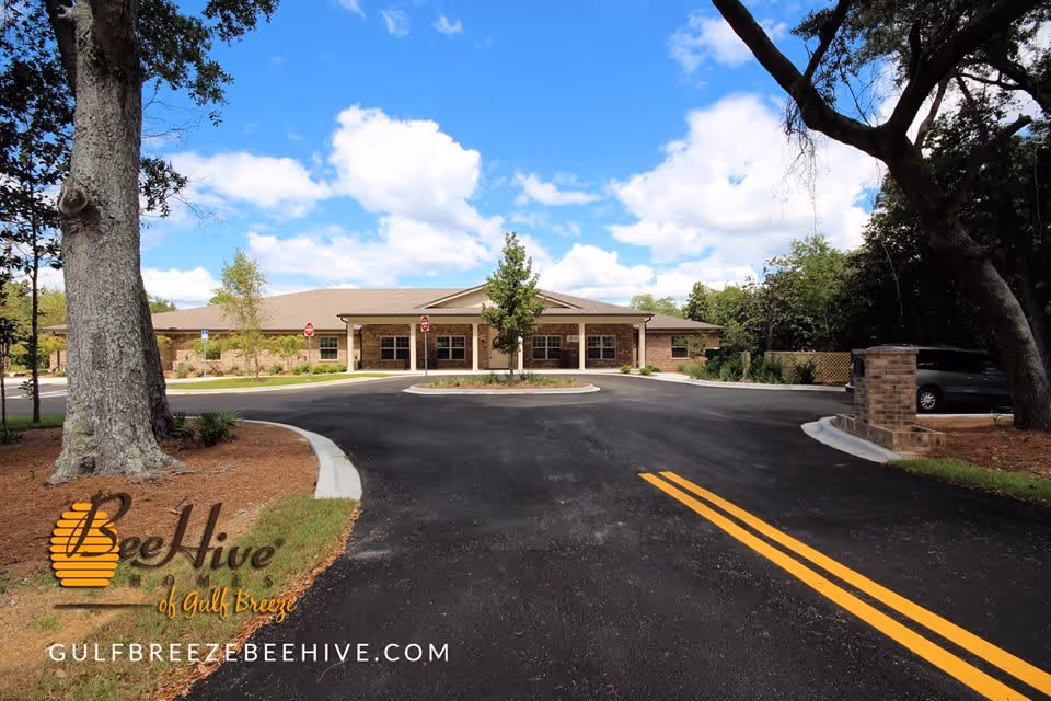Front exterior of a single-story BeeHive assisted living building with a round driveway, trees, and a blue sky.