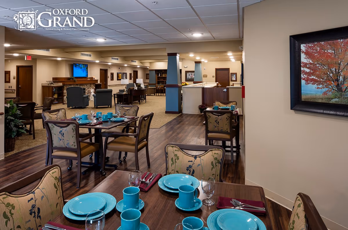 Interior view of a senior living facility dining area with several tables set with turquoise plates, cups, and silverware. The room has wooden flooring and beige walls, with a seating area featuring armchairs and a stone fireplace in the background. A framed picture of a tree with autumn leaves hangs on the wall to the right.