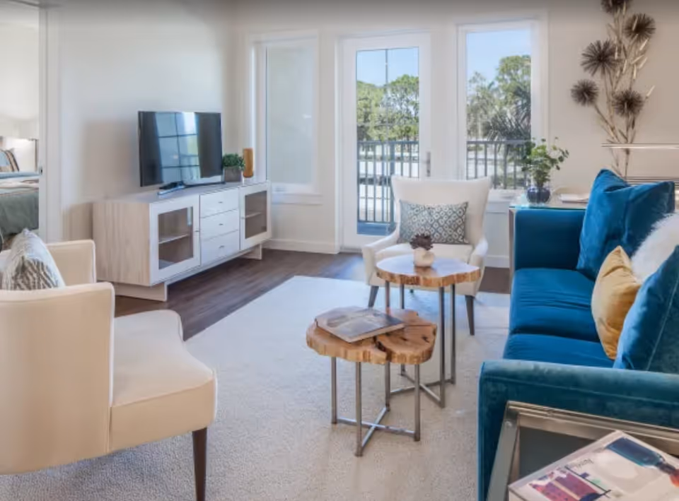 A bright and modern living room with a blue velvet sofa adorned with yellow and blue pillows, two white armchairs, and two wooden coffee tables with metal legs. A flat-screen TV sits on a light wood media console against the wall. Large windows and a glass door let in natural light and offer a view of greenery outside. The room has light-colored walls and a carpeted floor.