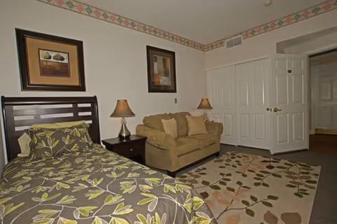 Furnished bedroom with a single bed, loveseat, nightstand lamps, framed artwork, and a patterned area rug.