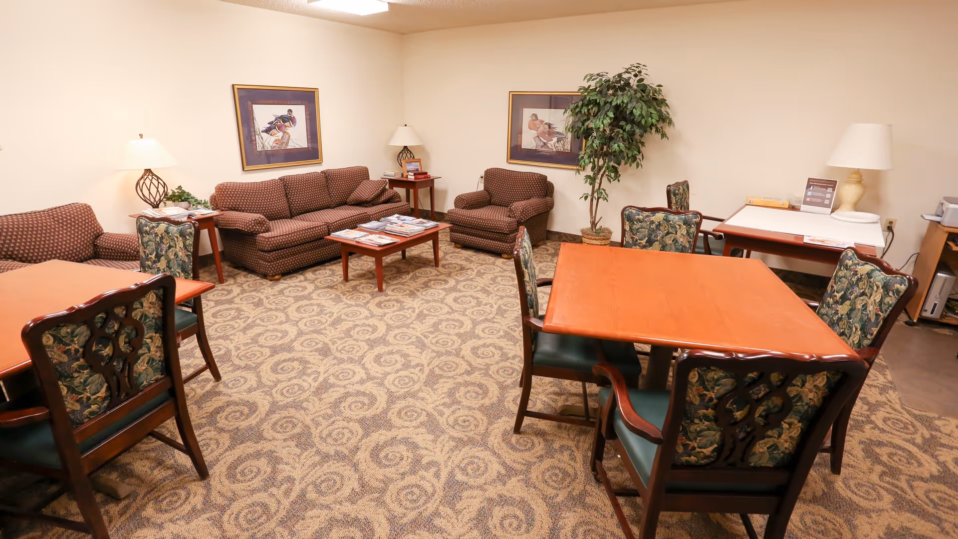 A cozy common room with patterned carpet, two wooden tables each surrounded by four floral upholstered chairs, two brown patterned sofas, an armchair, two framed bird paintings on the wall, two table lamps on side tables, a potted plant, and a small table with informational materials and a lamp in the corner.