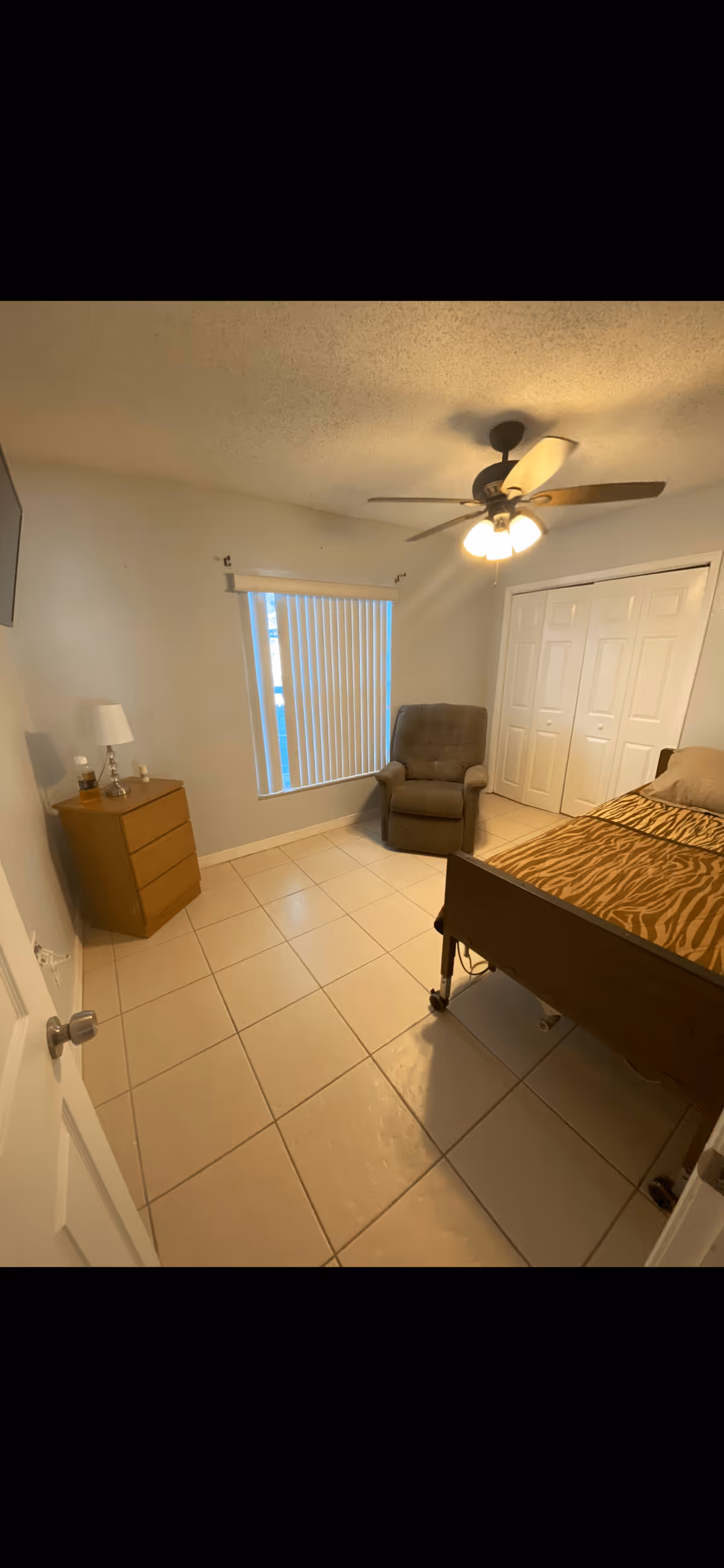 A small bedroom with beige tiled floor, a ceiling fan with lights, a window with vertical blinds, a brown recliner chair, a wooden dresser with a lamp and a bottle on it, and a bed with a tiger-striped blanket. There is a white closet with double doors on the far wall.