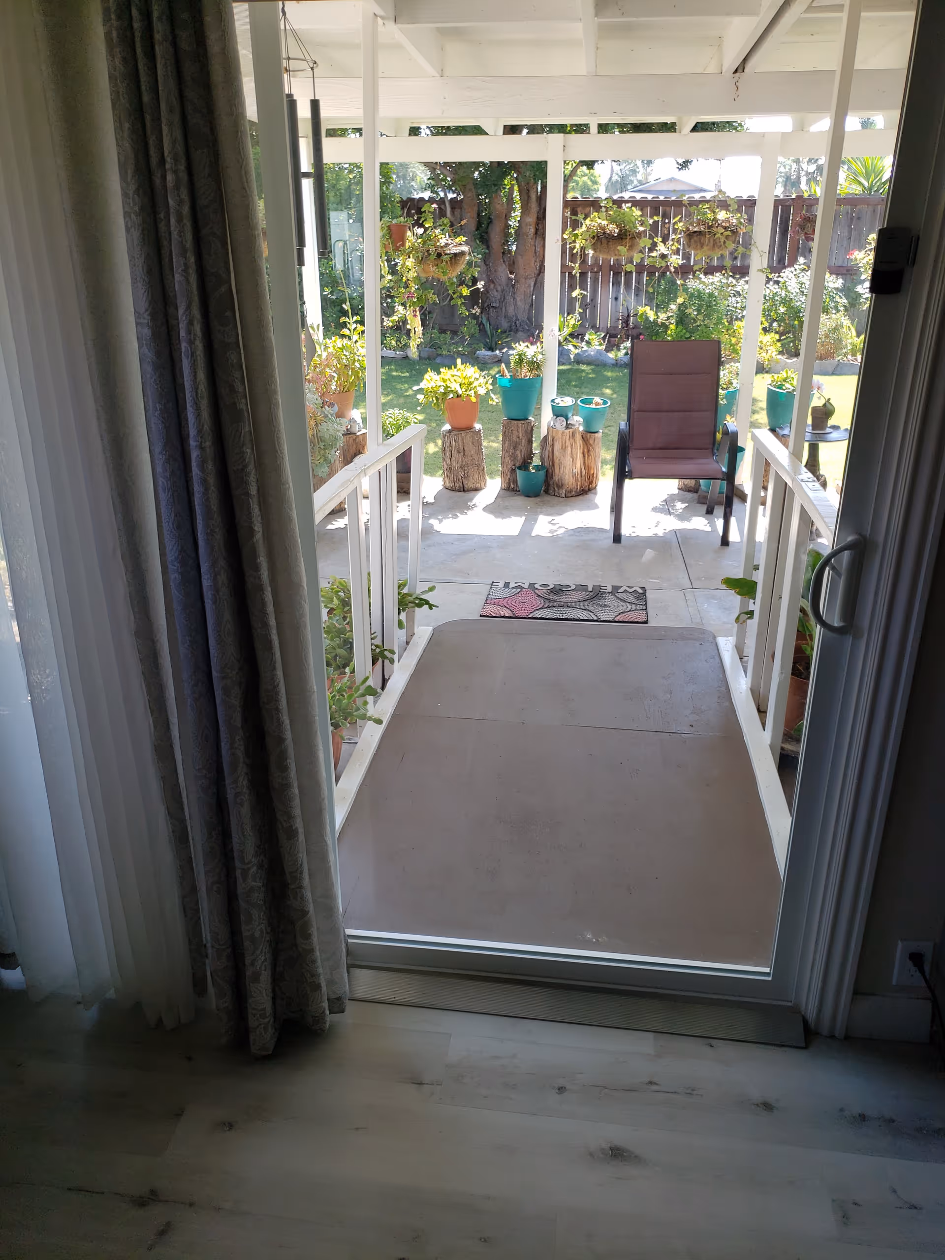 View through a glass sliding door from inside a room looking out onto a covered patio with a ramp leading down to a garden area. The patio has a brown chair, several potted plants on tree stump stands, and hanging plants. A welcome mat is placed at the bottom of the ramp.